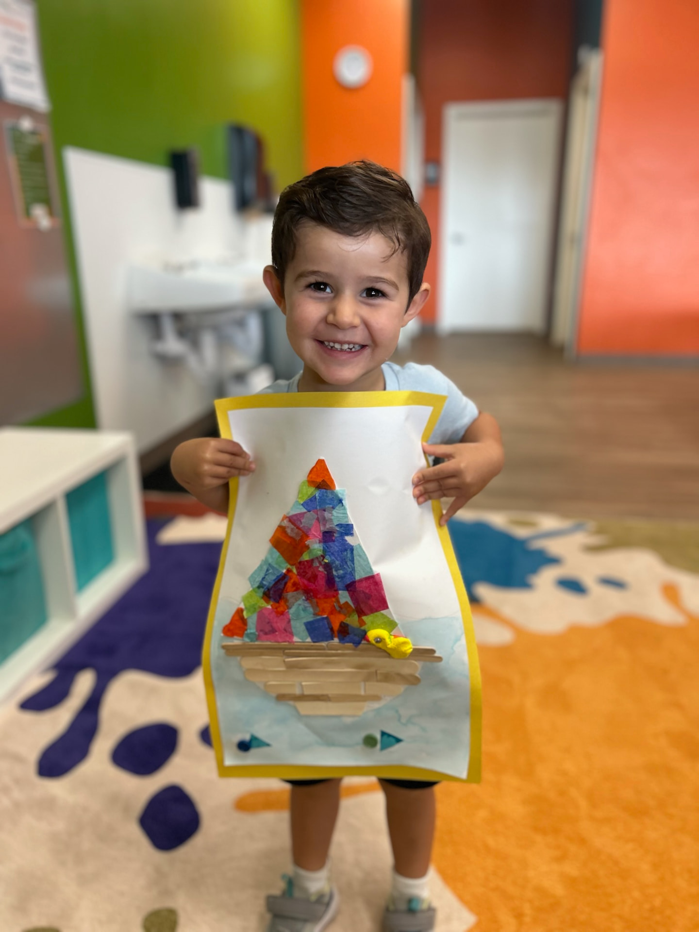 A boy smiling as he holds up a tissue-paper and popsicle-stick sailboat collage he made
