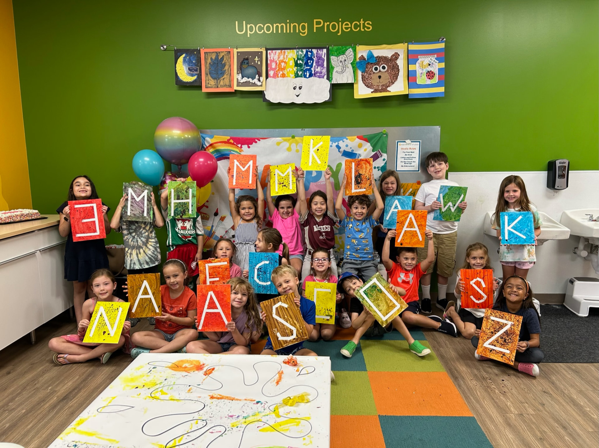Children on a studio rug holding up canvases they painted with letters of the alphabet